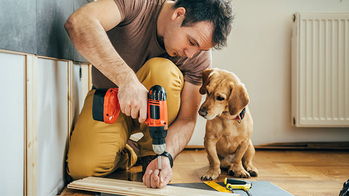 A man drilling into a wood plank while sitting next to a dog.